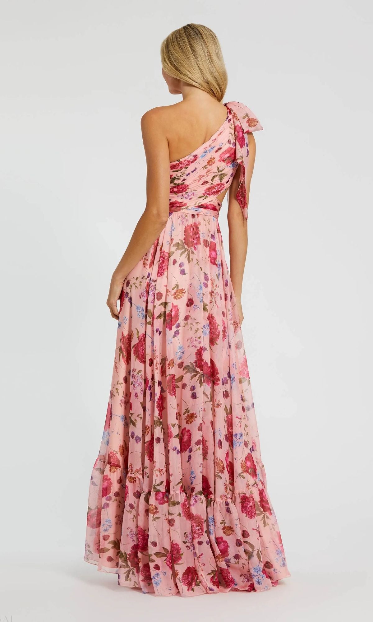 A woman faces away from the camera, showcasing the elegance of the Mac Duggal 68553— a pink, one-shoulder long formal dress with a shoulder bow and floor-length floral chiffon.